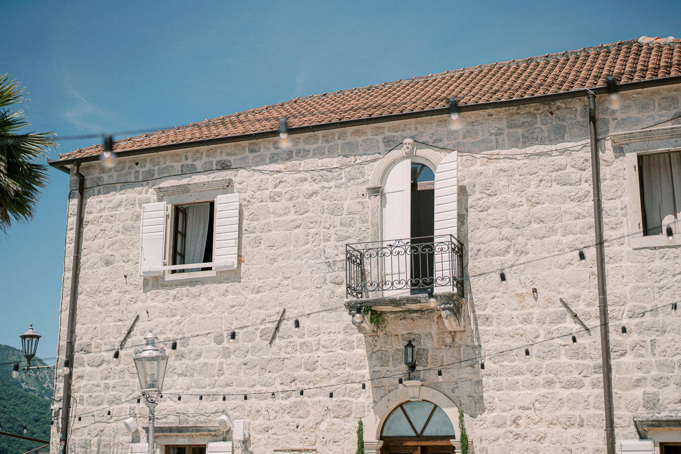Old stone building with open white shutters and a small wrought-iron balcony under a clear blue sky, surrounded by string lights and greenery.