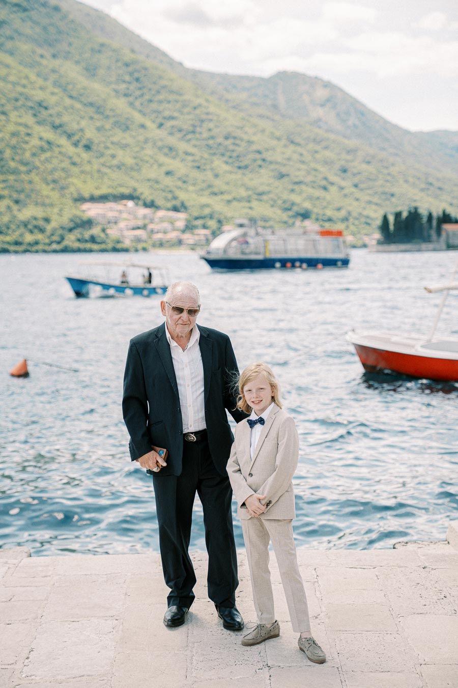 Elderly man and young boy in suits posing by the waterfront, with mountains and boats in the background on a sunny day.