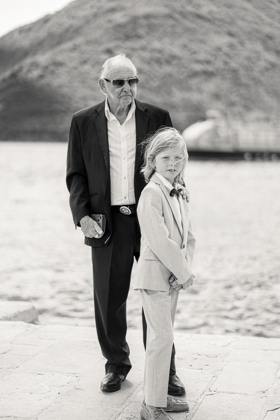 Black and white photo of an elderly man wearing sunglasses and a dark suit standing beside a young boy in a light suit and bow tie, outdoors with hills and water in the background.