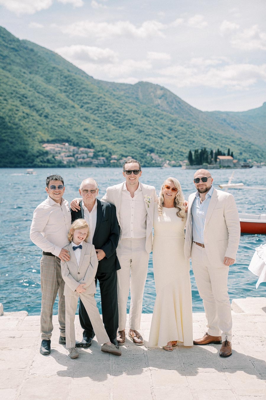 Family group photo by a scenic lake with mountains in the background, featuring men in suits and a woman in a dress on a sunny day.