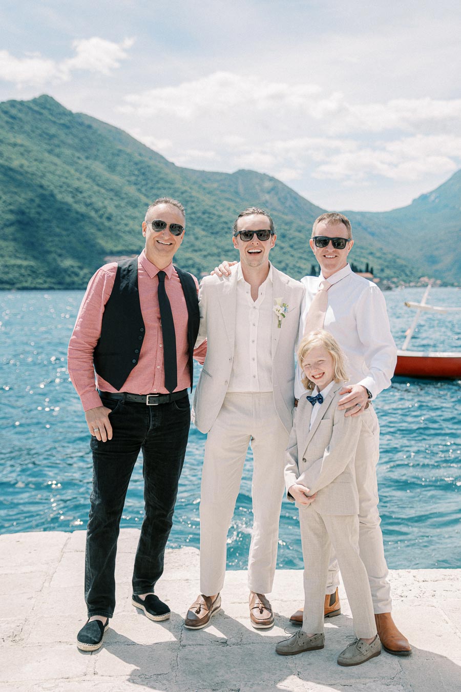 Group of four people in wedding attire smiling by a scenic waterfront with mountains in the background on a sunny day