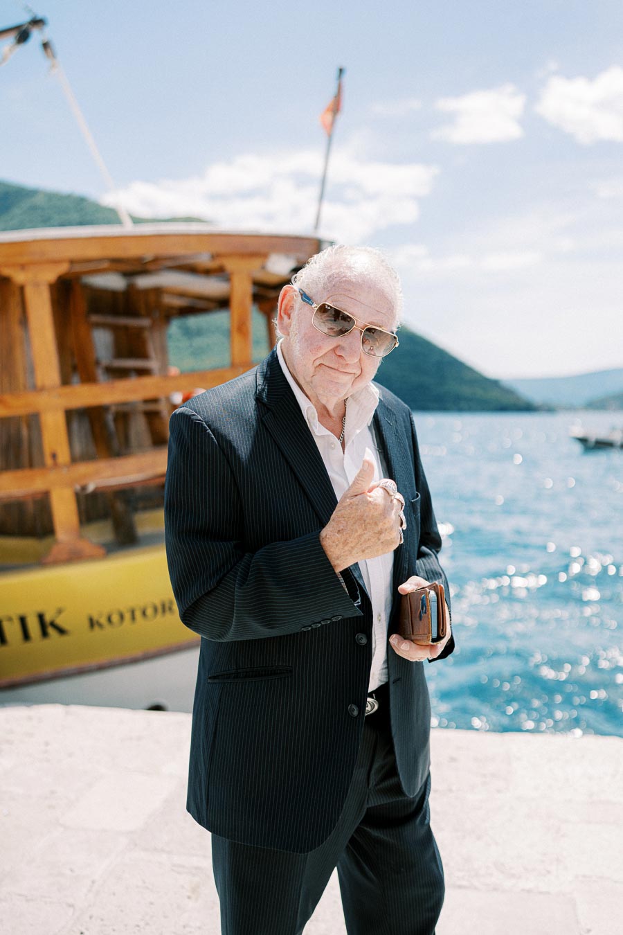 Elderly man in a stylish suit and sunglasses gives a thumbs-up by a scenic waterfront with a wooden boat and hills in the background.