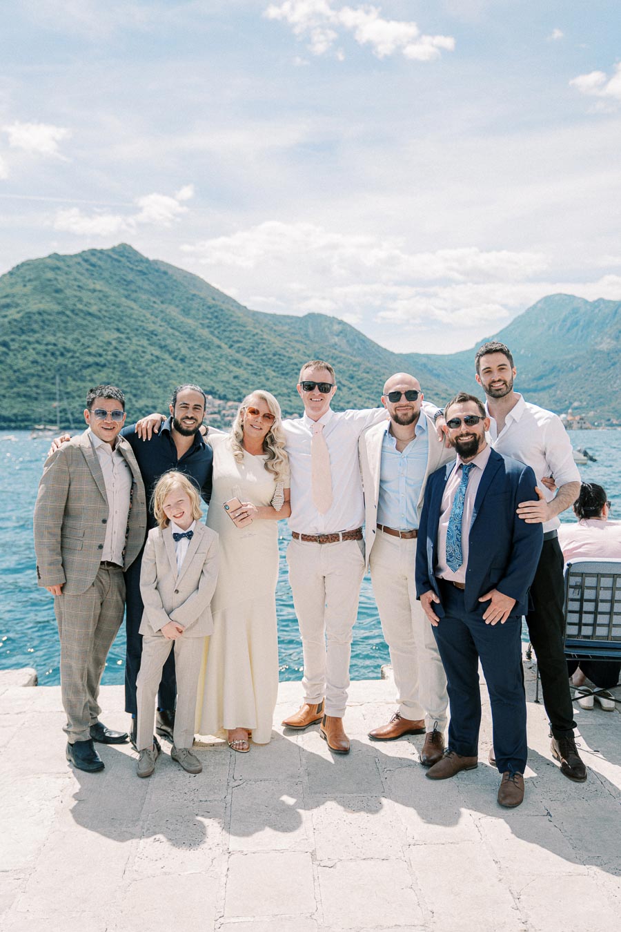 Group photo of elegantly dressed people at a scenic waterfront, with mountains and a bright blue sky in the background.