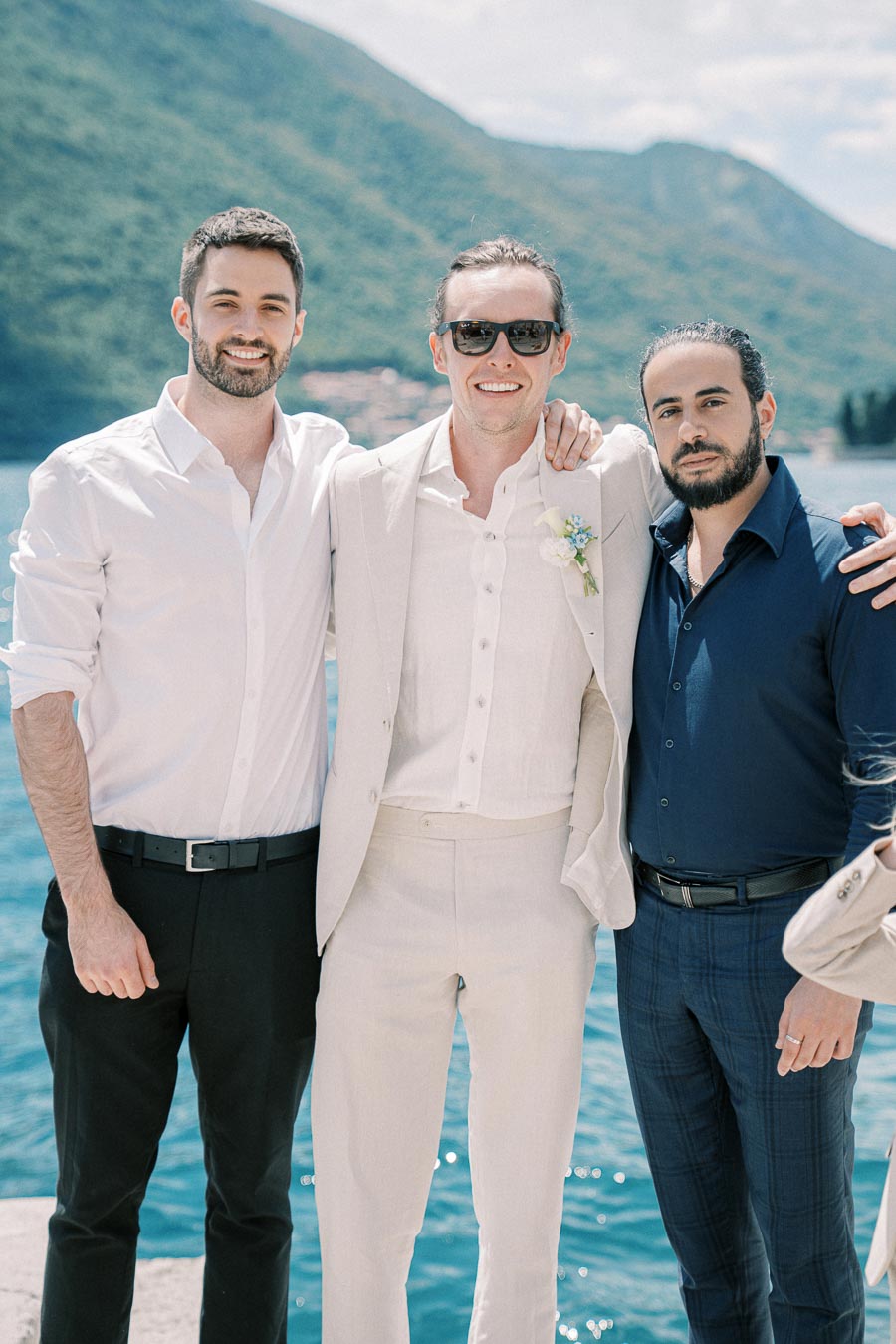 Three men in dress attire posing together by a scenic waterfront, with lush green mountains in the background.