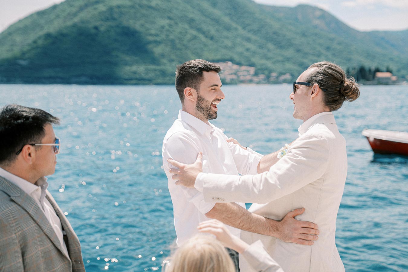 Three men on a boat congratulate each other with mountain and lake backdrop, wearing light summer attire under a clear blue sky.