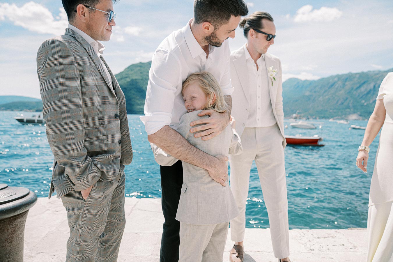 A joyful moment at a waterfront wedding, with a man in a white shirt hugging a child in a suit, surrounded by elegantly dressed guests; boats and mountains are visible in the background under a clear blue sky.