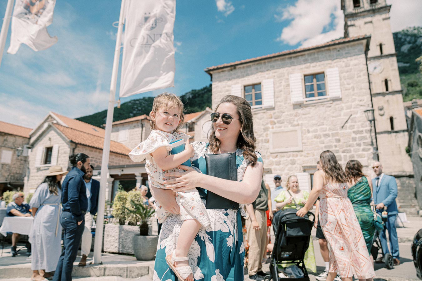 A woman in sunglasses holds a young girl outside a historic stone building on a sunny day, surrounded by people enjoying a gathering or event.