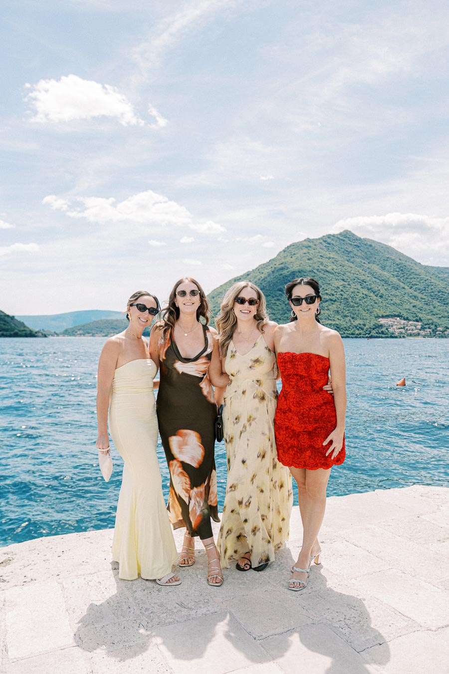 Four women in stylish dresses and sunglasses standing together by a scenic lakeside with mountains in the background under a sunny blue sky.