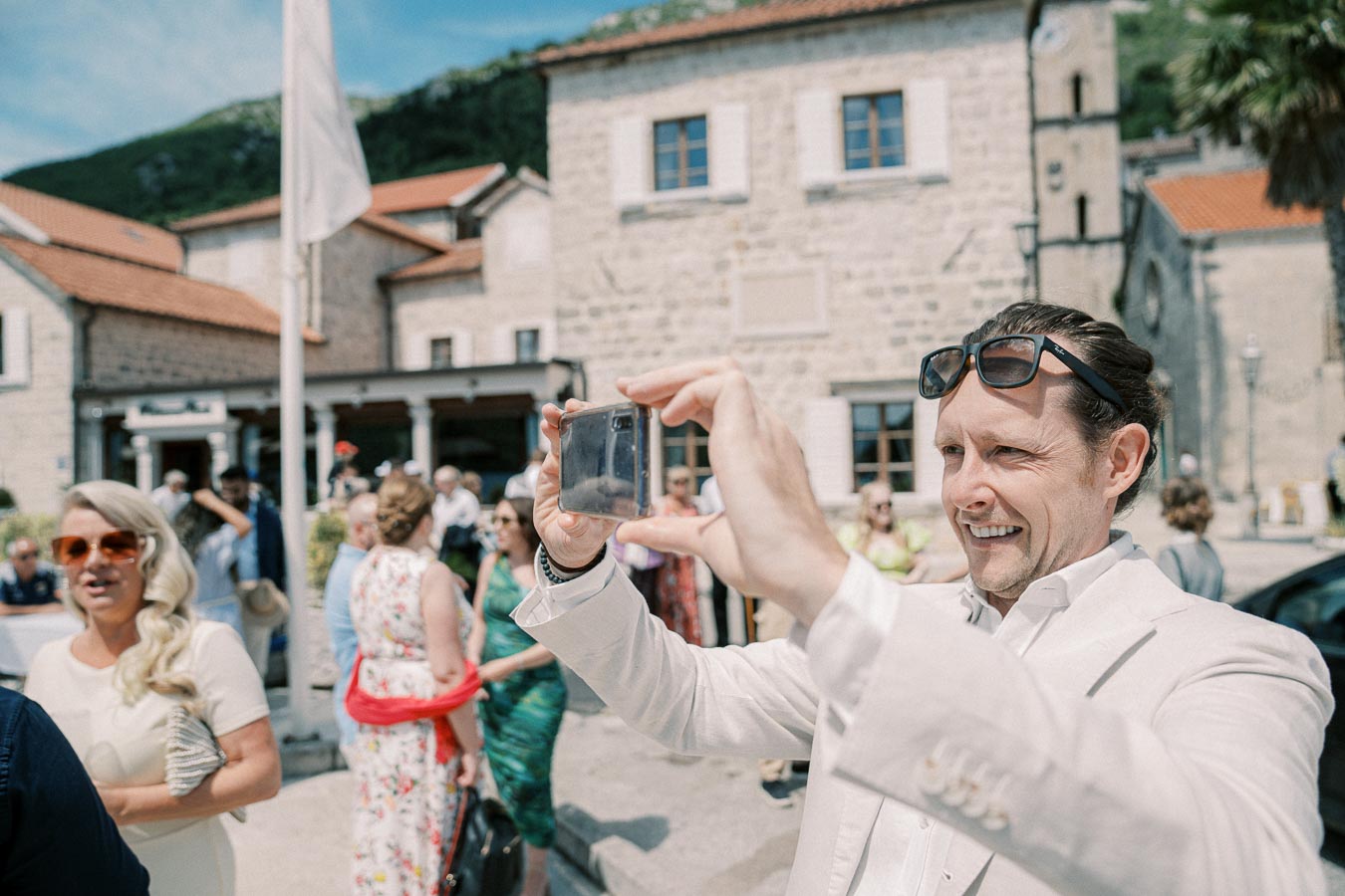 A man in a light suit and sunglasses is smiling while taking a photo with a smartphone during an outdoor social event, with a group of people and a historic stone building in the background.