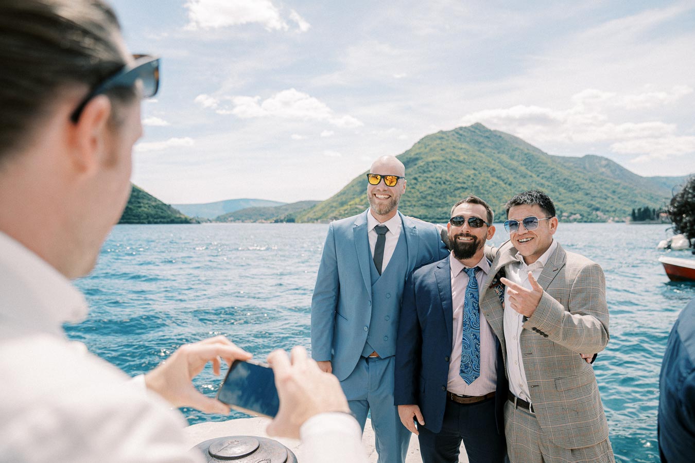 Three men in suits and sunglasses pose for a photo on a boat with mountains and a lake in the background, while another person takes their picture.