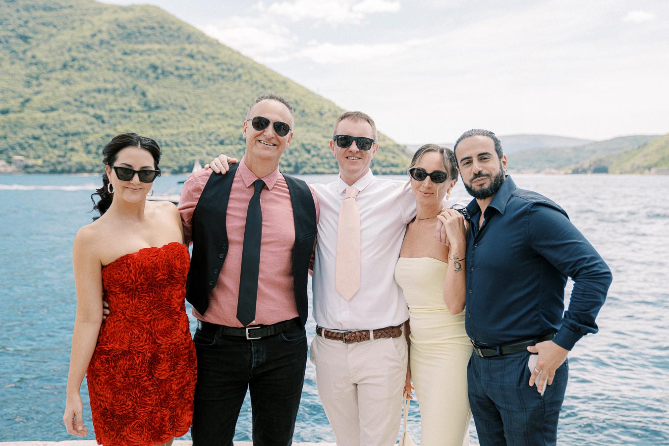 Group of five people in stylish summer attire posing happily in front of a scenic lake and mountain backdrop.