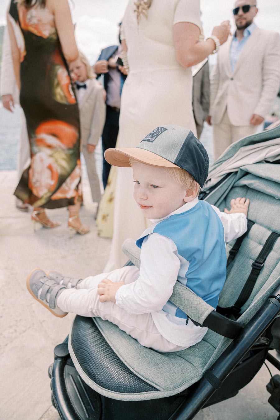 A young child wearing a blue vest and a cap sits in a stroller during an outdoor gathering, surrounded by elegantly dressed adults in light-colored attire.
