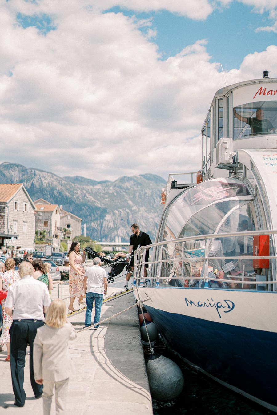 Tourists boarding a boat named 'Marija D' at a scenic coastal port with mountainous backdrop under a bright blue sky, capturing a vibrant travel scene.