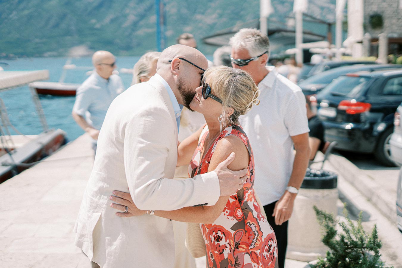 A couple exchanging a kiss at an outdoor summer gathering by a marina, surrounded by people and parked cars, with scenic mountains and a lake in the background.