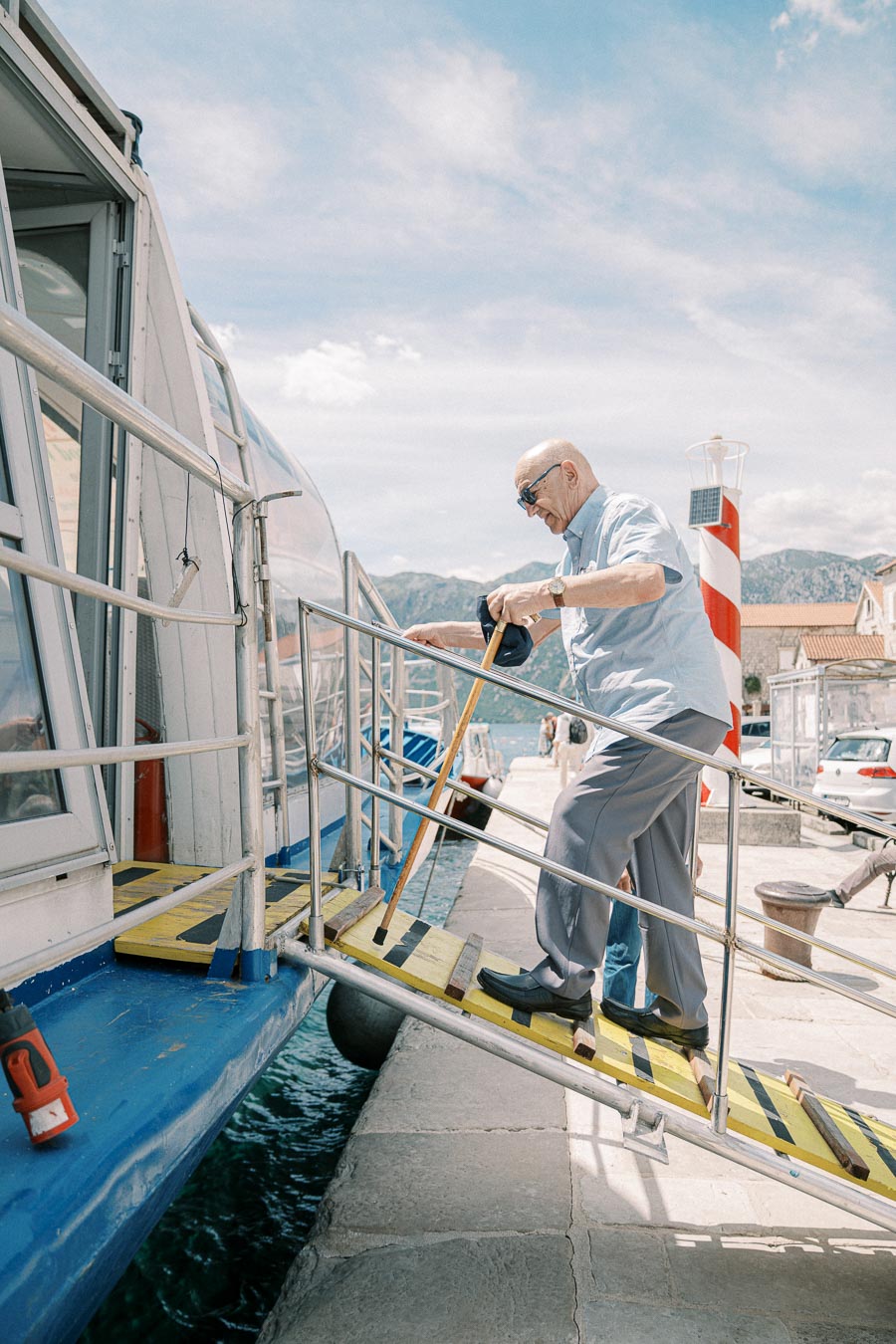 Elderly man with a walking cane boarding a boat using a ramp on a sunny day at a harbor with a lighthouse in the background.