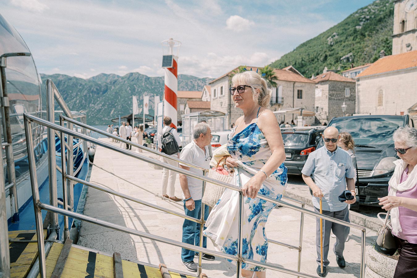 Elderly woman in a floral dress boarding a boat on a sunny day at a picturesque harbor with mountains in the background.