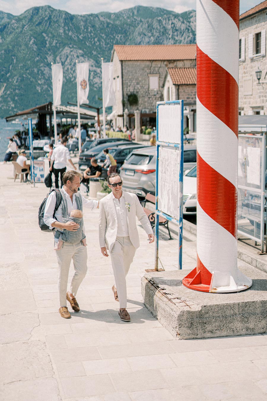 Two men walking beside a red and white striped pole on a picturesque waterfront promenade, with mountains and historic buildings in the background, under a clear sky.