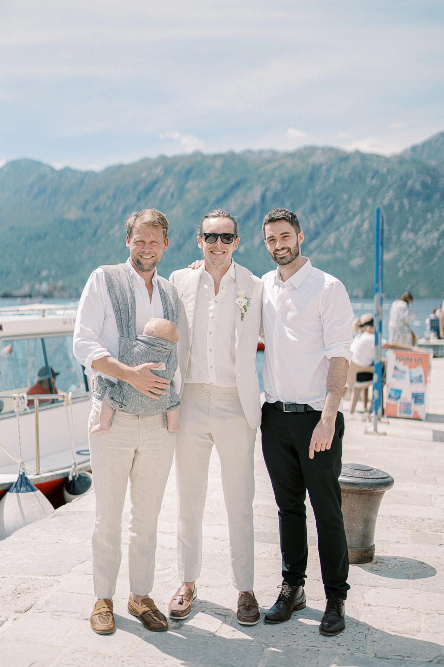 Three men pose happily by a mountainous waterfront, with one man carrying a baby in a sling. The scene captures a casual, sunny day by the harbor with boats in the background.