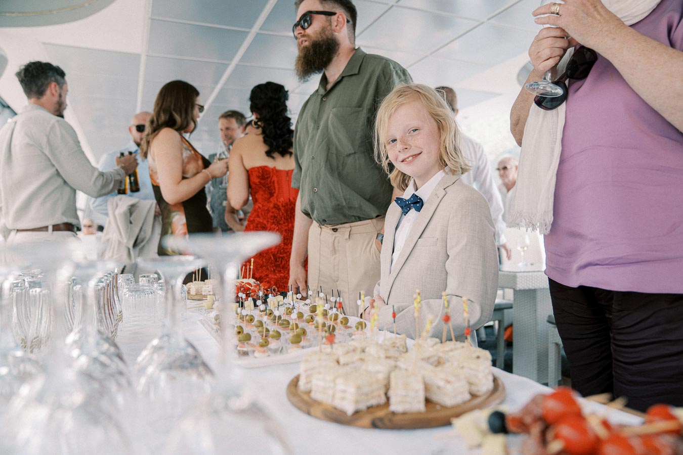 A group of people at a social event on a yacht with a well-dressed child smiling at the camera. The table in the foreground is elegantly set with appetizers and empty glasses, capturing a festive atmosphere.