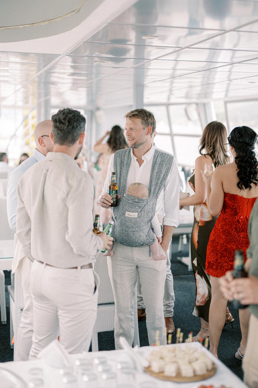 A group of people socializing at a party on a boat, with one man holding a baby in a carrier and a drink, wearing light-colored clothing. The setting is casual and includes a mix of men and women.