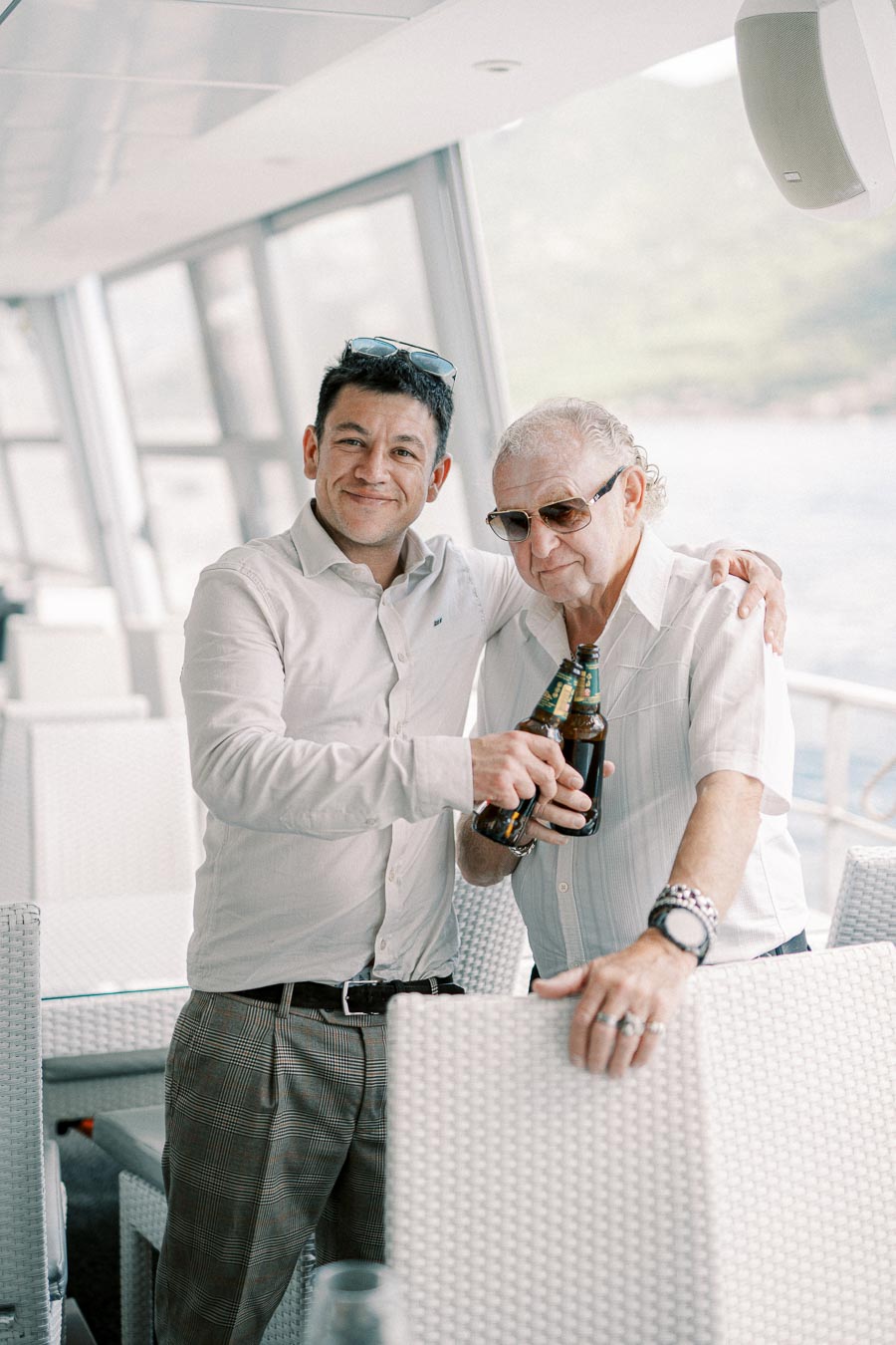 Two men enjoying a boat ride, clinking beer bottles and smiling at the camera, with a scenic water view in the background.