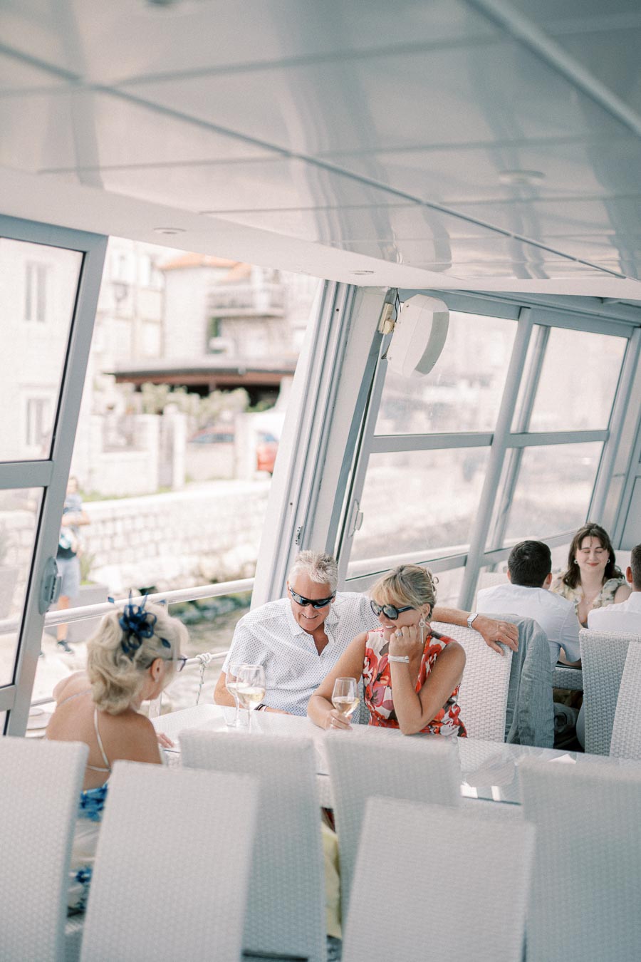 A group of people enjoying a relaxing meal and conversation on a sunny patio with large windows overlooking a scenic view.