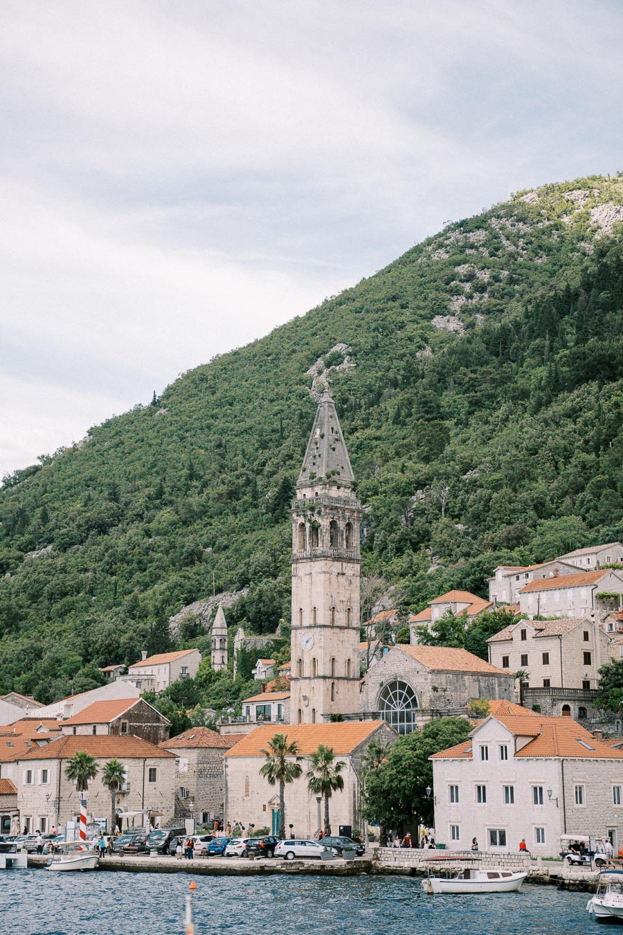 Scenic view of a historic coastal town with a tall stone bell tower surrounded by green hills, featuring charming stone buildings with orange-tiled roofs and boats docked by the waterfront.