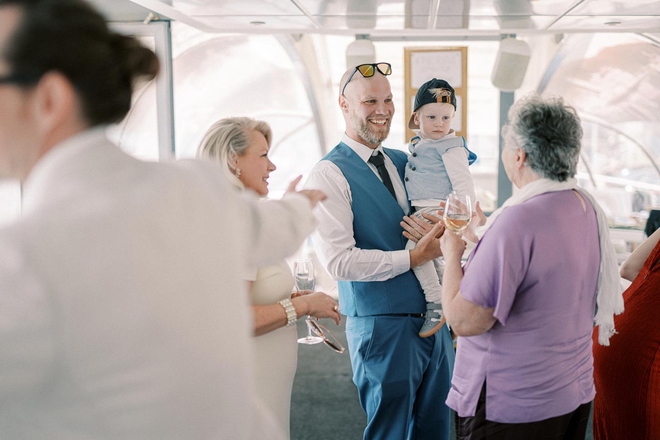 A group of people enjoying a social gathering indoors, with a man in a blue vest holding a toddler. They are smiling and interacting, with drinks in hand, creating a warm and friendly atmosphere.