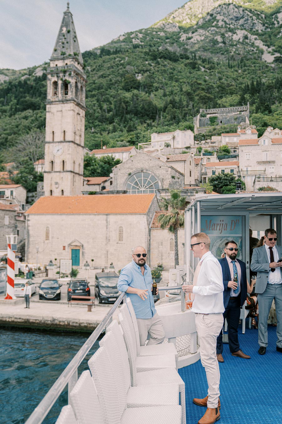 Group of men socializing on a boat near the historic Perast bell tower and church, set against a backdrop of lush mountains in Montenegro.