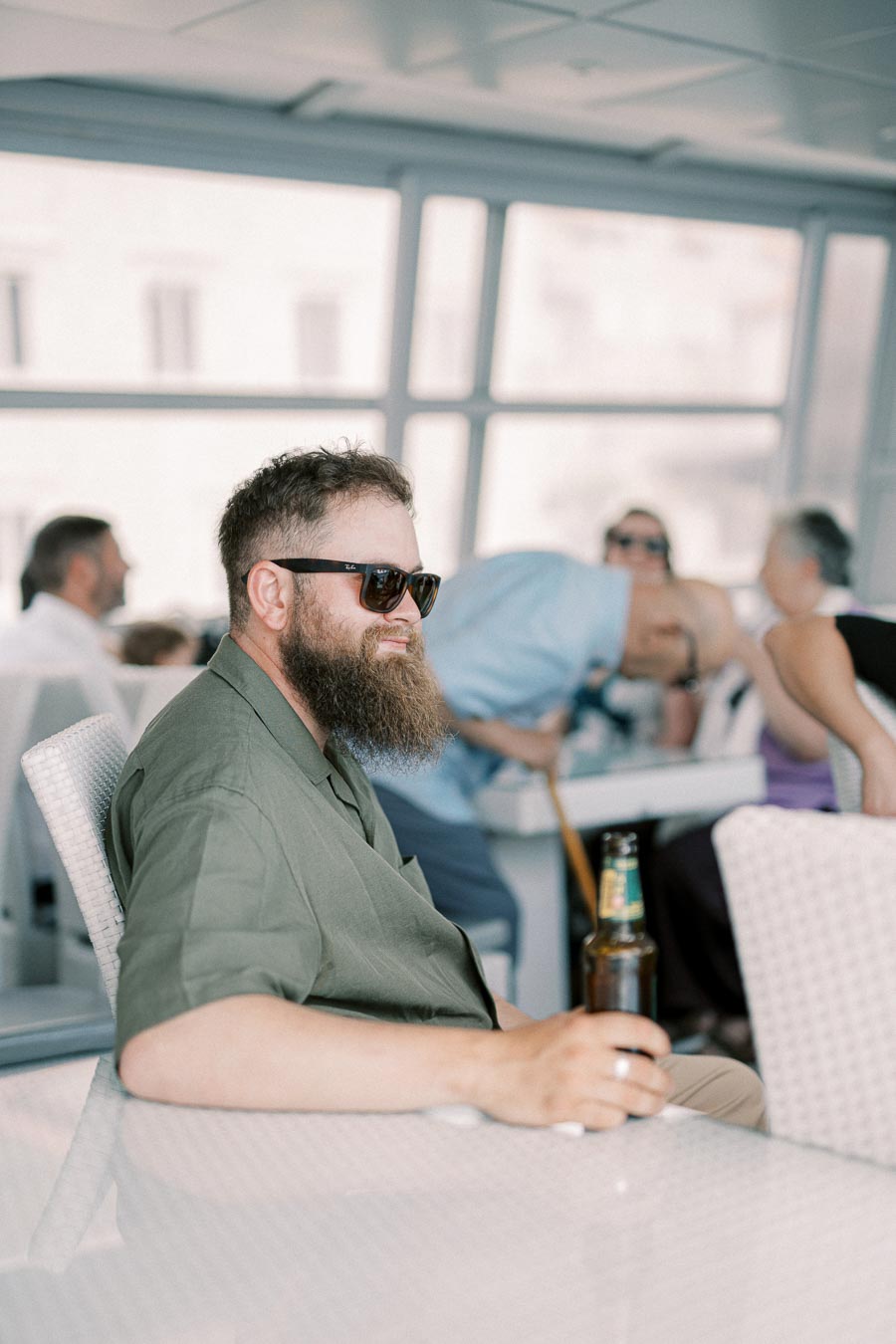 Man with a beard and sunglasses sitting indoors, holding a beer bottle in a relaxed setting.