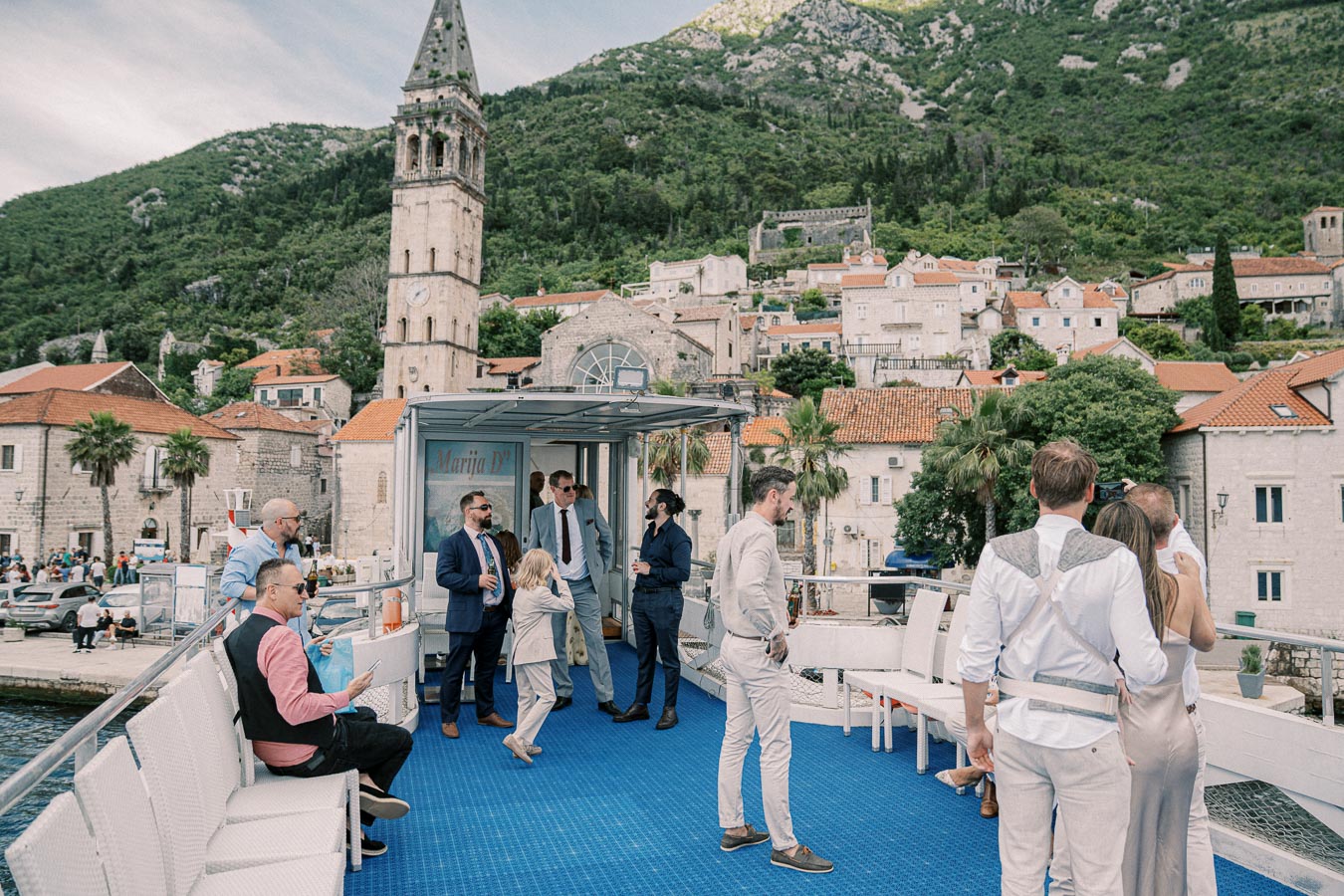 A group of well-dressed people socializing on a boat, with a historic bell tower and scenic hillside village in the background. Perfect setting for a wedding or event by the waterfront.