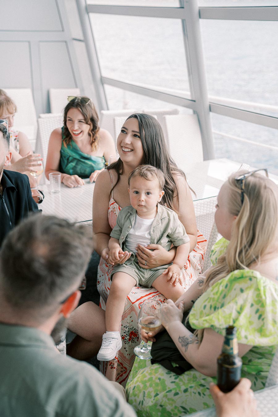 A joyful group of people enjoying a gathering on a boat, with a woman holding a toddler on her lap. The setting features large windows with a view of the water, creating a relaxed and happy atmosphere.