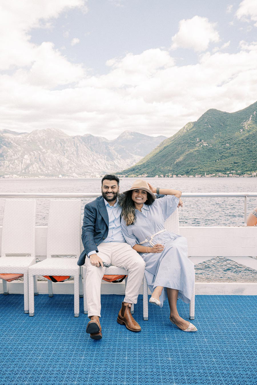 A couple smiling on a boat with a scenic backdrop of mountains and lake, under a partly cloudy sky.