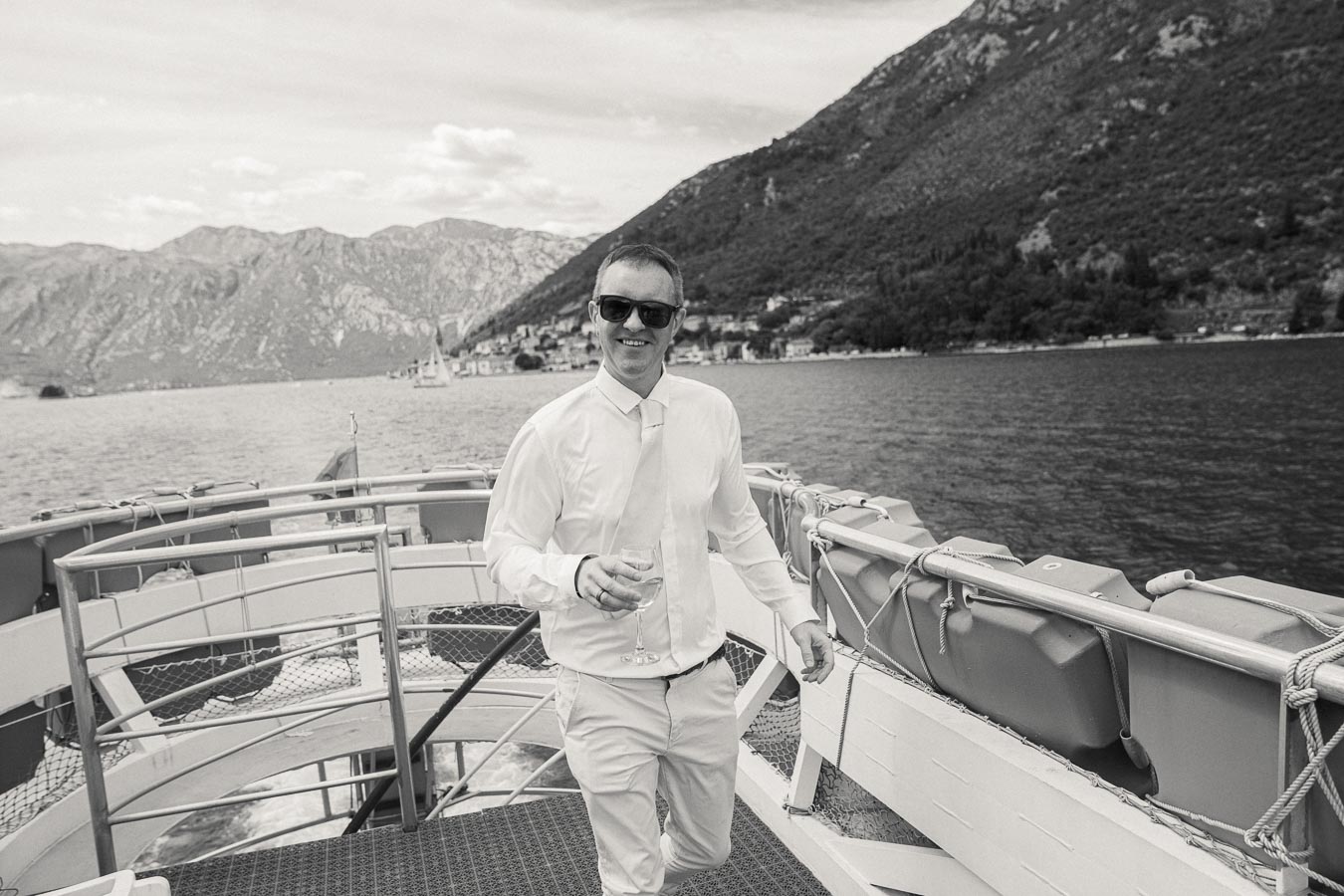 A man in sunglasses and formal attire holding a glass, standing on a boat with scenic mountain and sea views in the background.