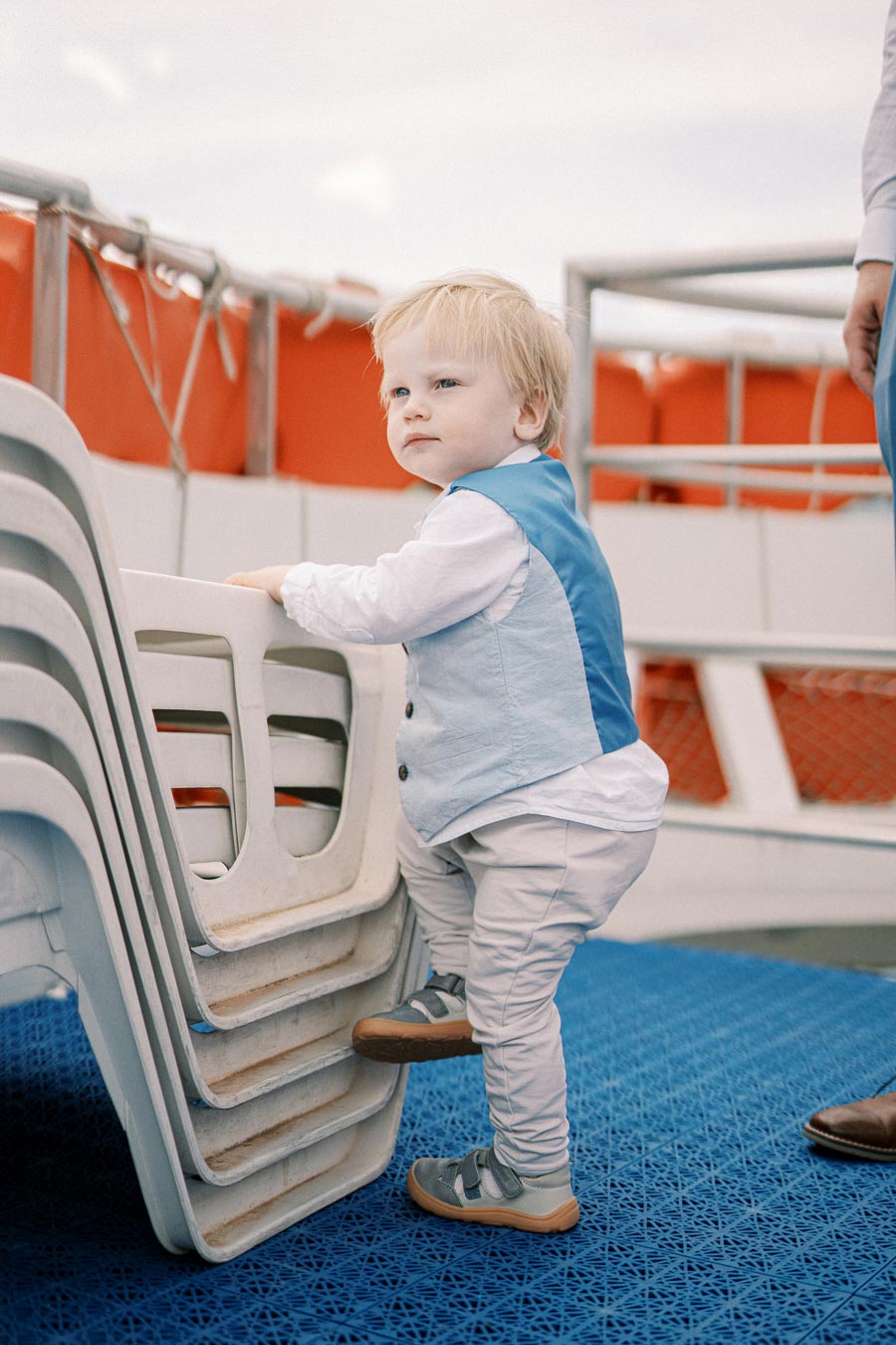 Toddler in stylish outfit leaning on stacked chairs aboard a boat, with blue flooring and orange safety gear in the background.