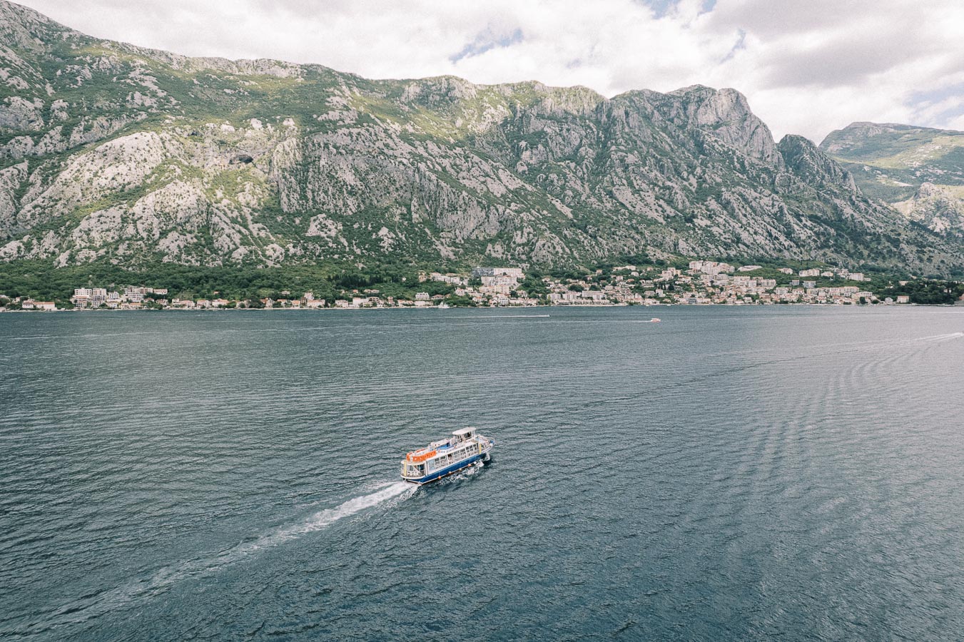 A scenic view of a boat cruising on a tranquil bay with mountainous terrain in the background, under a partly cloudy sky.