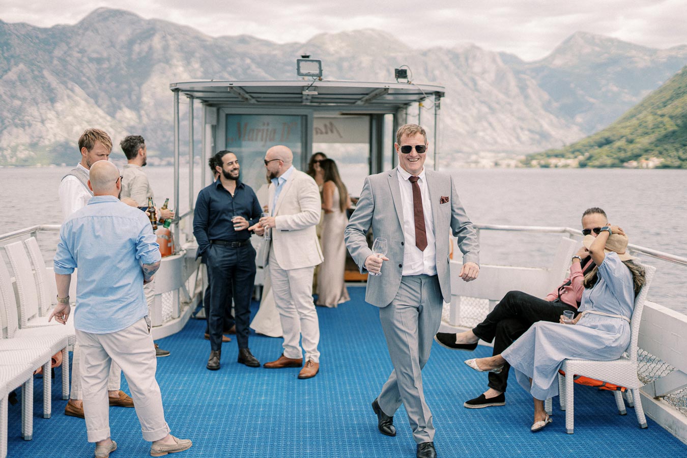 A vibrant group of people enjoying a social event on a boat with scenic mountain views in the background, featuring a man in a gray suit walking confidently, holding a glass, surrounded by others in casual and formal attire.
