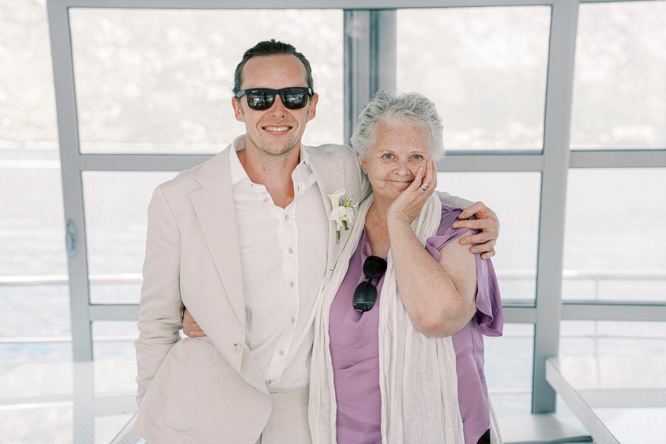 A stylish man in a light suit, wearing sunglasses, posing with an elderly woman in a purple top, smiling inside a modern building by the water.