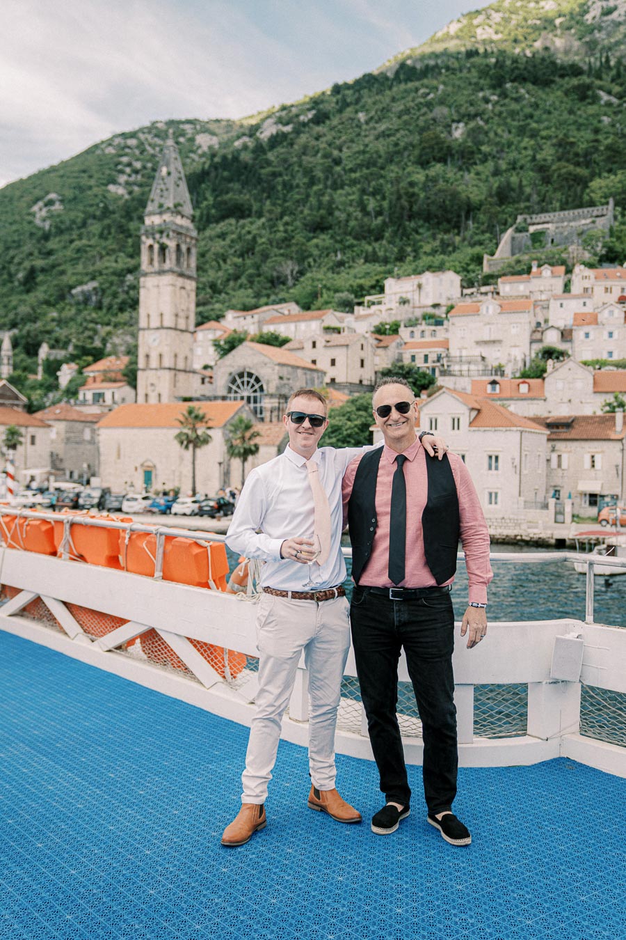 Two people wearing sunglasses and formal attire pose together on a boat, with a picturesque historic town and lush green hills in the background.