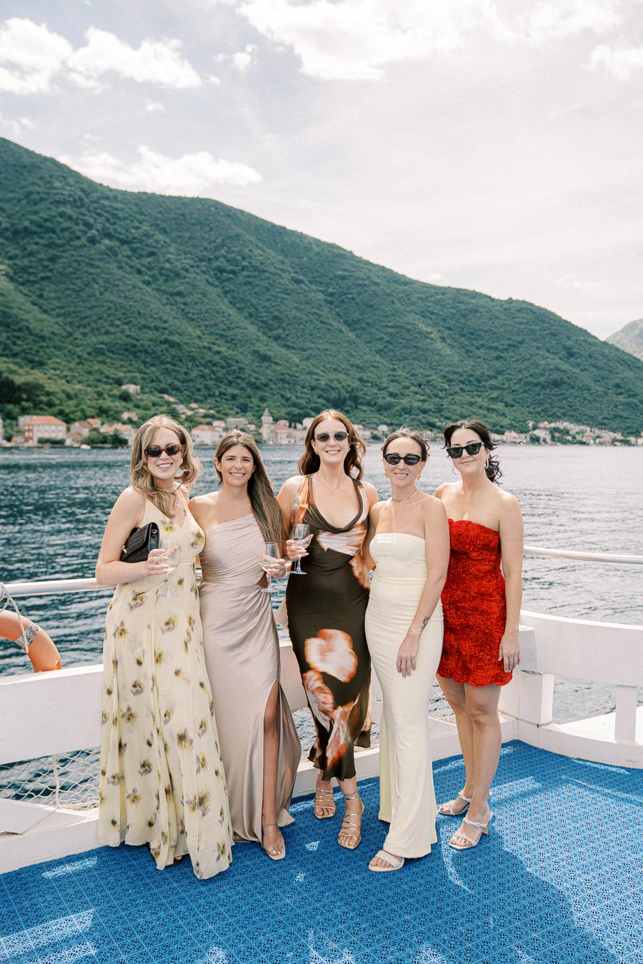 Five women in elegant dresses, holding wine glasses, stand on a boat with a scenic view of lush green mountains and a tranquil lake in the background under a partly cloudy sky.