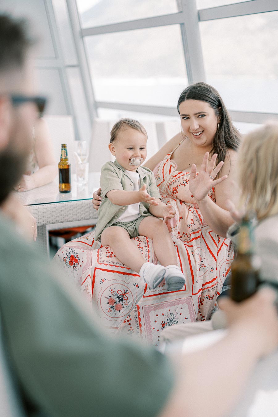 A woman in a floral dress holding a baby with a pacifier, sitting at a glass table with drinks, engaging in conversation, indoors with a scenic view.