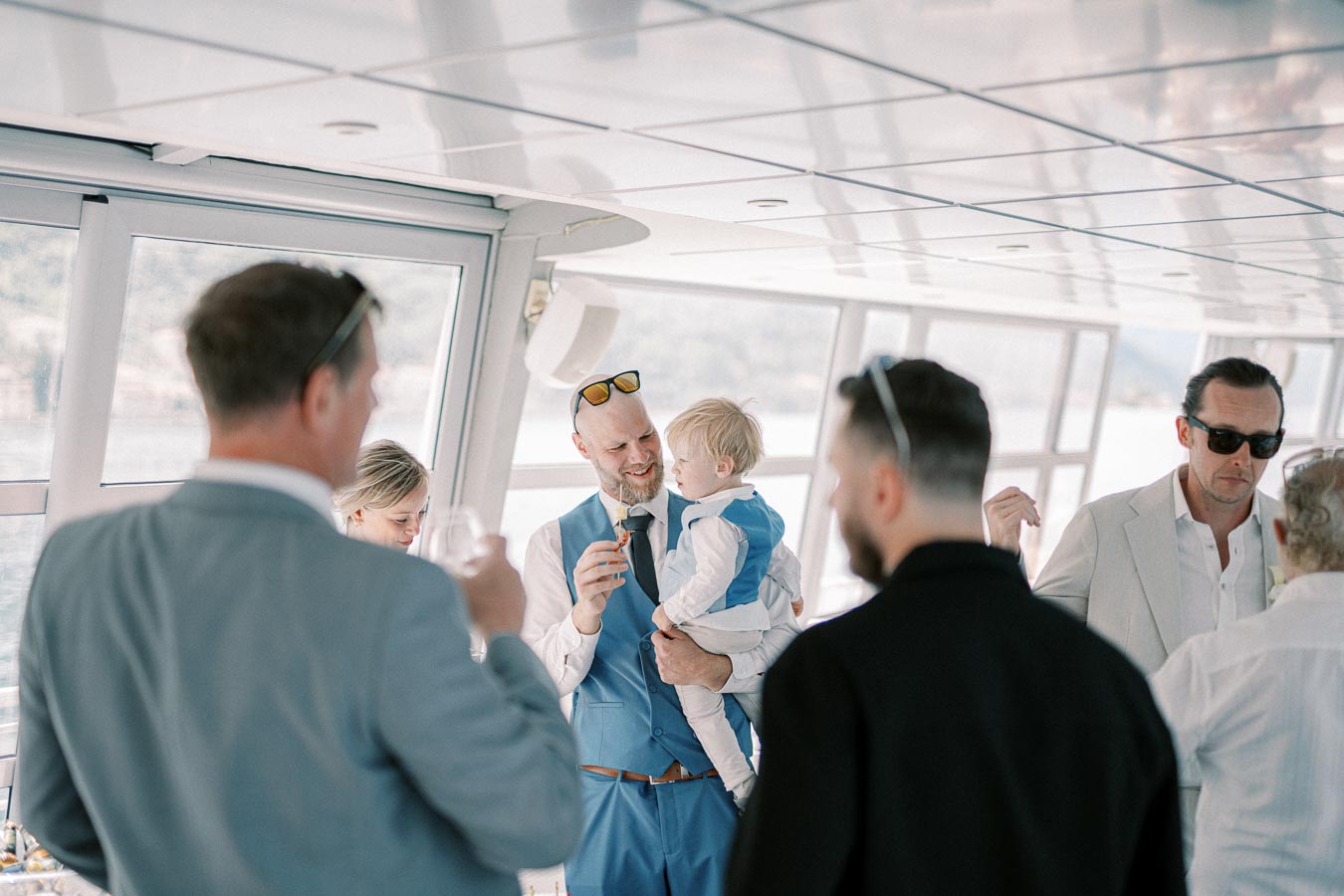 A group of people enjoys a formal gathering on a stylish boat, with a man in a blue suit smiling and holding a toddler dressed in matching attire, creating a joyful atmosphere.