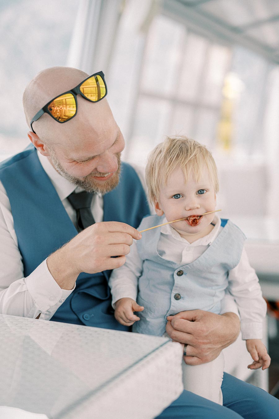 Father and toddler son bonding, both dressed in blue vests, as the father playfully feeds the child a treat on a skewer.