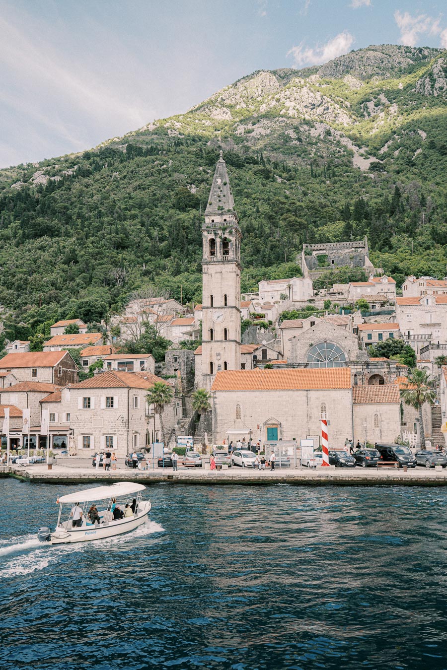 Scenic view of Perast, Montenegro, featuring historic church architecture with a prominent bell tower, surrounded by picturesque mountains and waters, with a boat gliding across the bay.