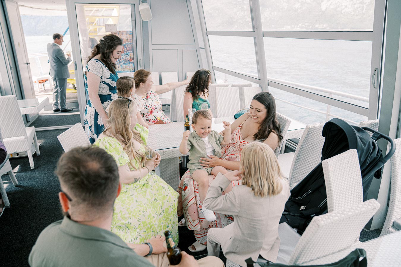 A group of people enjoying a social gathering on a boat, with a scenic view through large windows. A woman in a floral dress smiles while holding a young child.