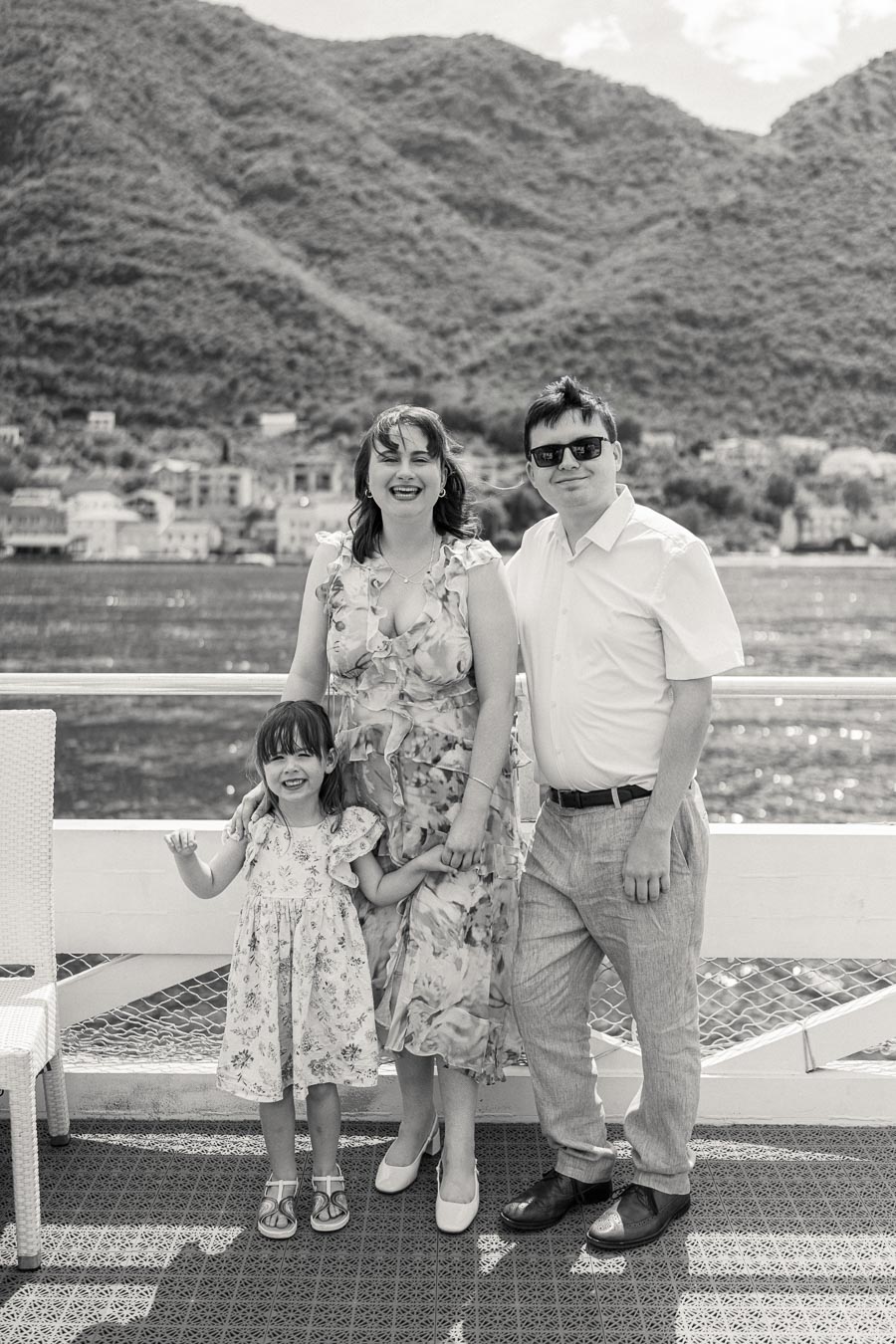 Family enjoying a scenic view by the water with mountains in the background, smiling and dressed in summer clothing, black and white photography.