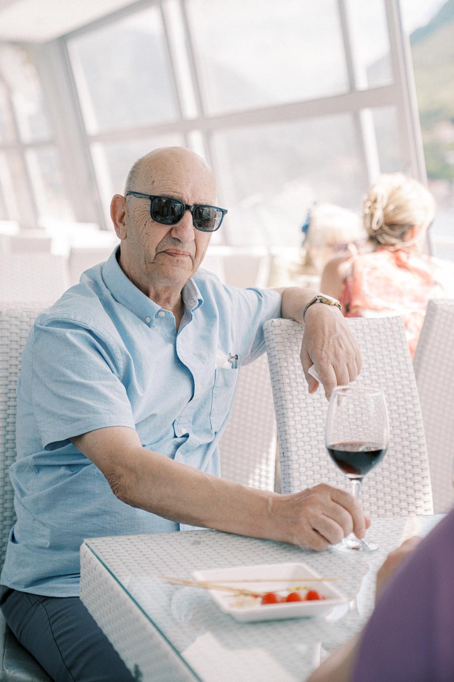 Elderly man wearing sunglasses, sitting at an outdoor cafe table with a glass of red wine and small snack plate, enjoying a sunny day.