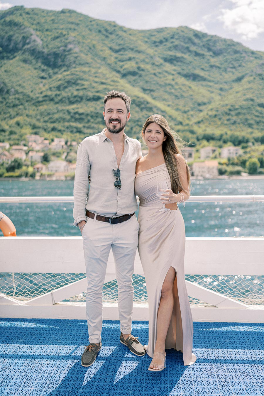 A couple standing on a boat with scenic green mountains and a blue lake in the background, dressed elegantly for a day out on the water.