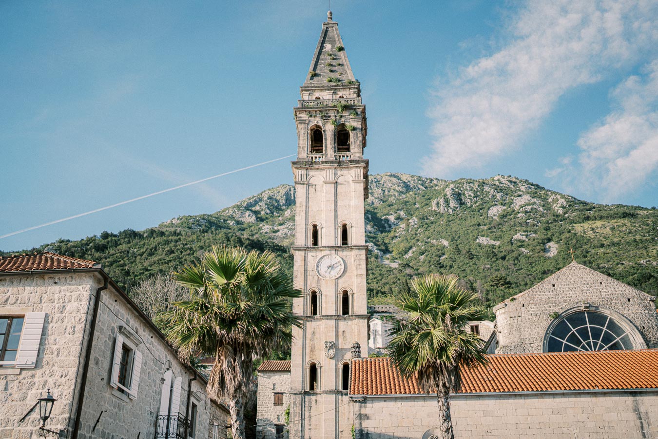 Ancient stone clock tower in a historic Mediterranean town with palm trees and mountain backdrop under a clear blue sky.