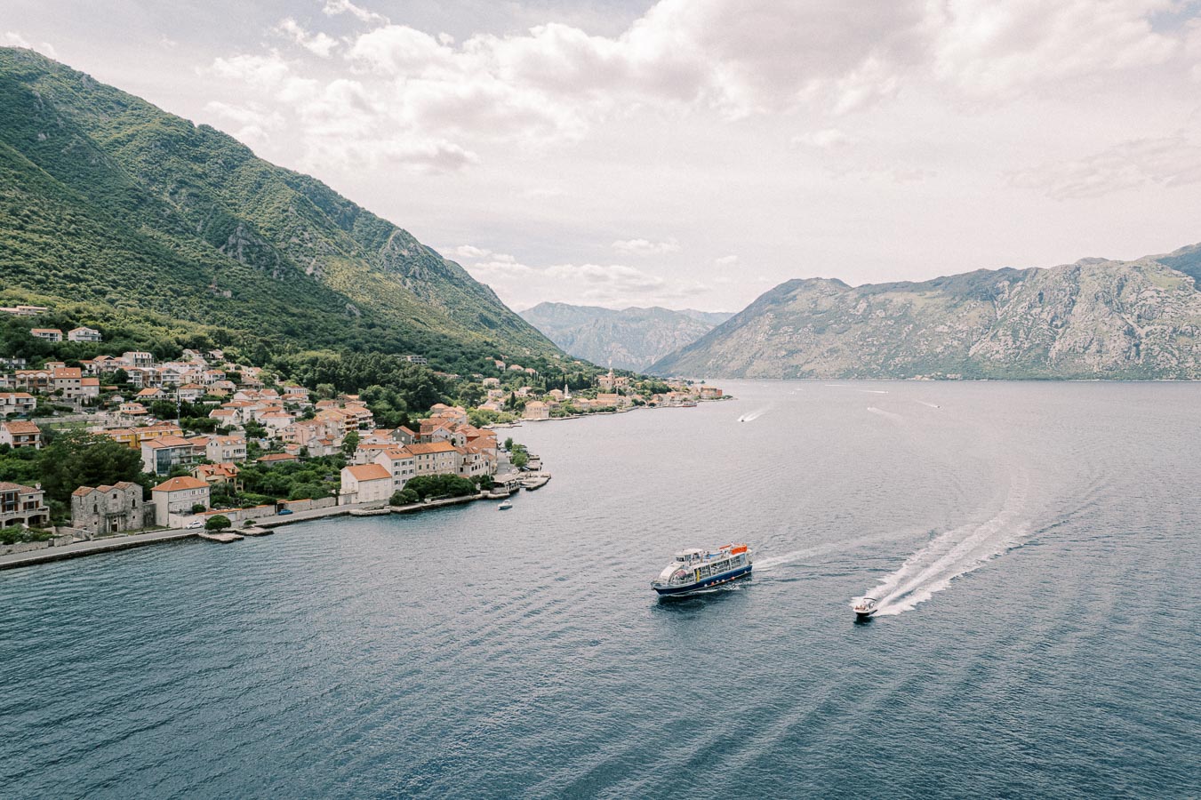 Aerial view of a scenic coastal town with terracotta-roofed buildings nestled along the shoreline, surrounded by lush green mountains and a calm blue sea with a ferry and small boat traversing the water, under a partly cloudy sky.
